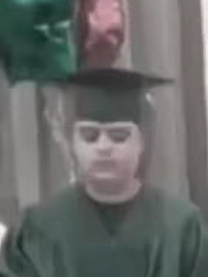 A person in a graduation cap and gown is the main subject. The photo is slightly out of focus, with the person's face visible. In the background, there are hints of other people and possibly some balloons.