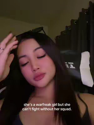 A medium shot shows a young woman, who is the main subject as she is positioned in the front of the image. She is Asian with long dark hair framing her face but parted to the side. She is wearing sunglasses on top of her head. She is gazing downward with a soft expression. The text overlay at the bottom of the frame indicates that she can't fight without her squad. The background is dark, suggesting a dimly lit indoor setting, and a gaming chair is visible.