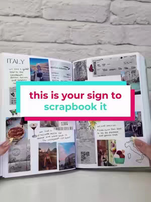 An open scrapbook is displayed, filled with photos and ephemera from a trip to Italy. A large text overlay reads 'this is your sign to scrapbook it'. The background is a white brick wall.