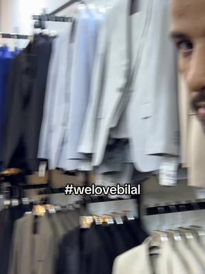 A man partially visible on the right side of the frame stands in what appears to be a clothing store, looking towards a wall of suits. The suits are of various colors and styles, hanging from racks. The lighting is bright, highlighting the formal wear.
