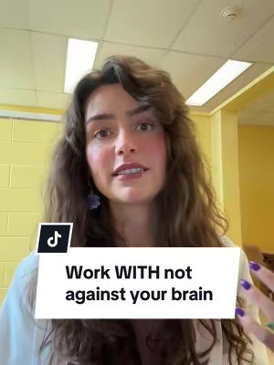 A medium shot captures a woman with long, wavy brown hair looking directly at the camera. She has a fair complexion and is wearing a light-colored top. A text overlay in a white box at the bottom of the frame reads, "Work WITH not against your brain."