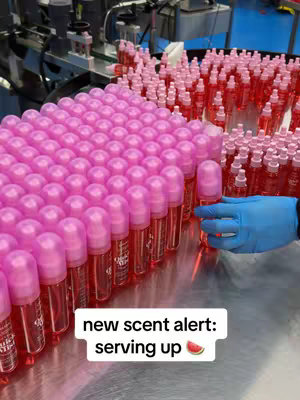 A close-up product shot featuring rows of perfume bottles on a metallic surface. Each bottle is filled with a red liquid and topped with a pink cap. A gloved hand selects one of the bottles. The background shows machinery.