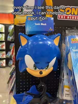 The image captures a display in a store, with a large close-up of a Sonic the Hedgehog mask hanging on a black grid. Above the mask, a sign reads "Sonic Mask." Text overlayed on the image says, "every time I see this damn sonic mask, i can't help but put it on." The overall composition is well-lit with focus on the Sonic mask.