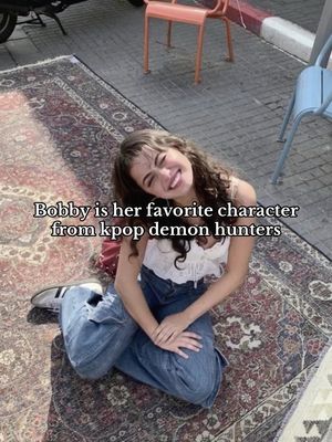 The cover image features a young woman sitting on a patterned rug, smiling at the camera. The background includes outdoor elements such as chairs and an exterior surface. Text is overlaid, describing the woman's favorite character.