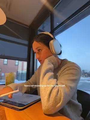 A woman sits at a table near a window, facing left, wearing headphones and looking at her tablet. She is in a cafe setting.