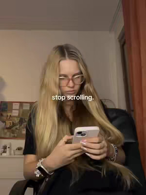 A young woman with long blonde hair and glasses is sitting in a chair and looking at her phone. The text overlay "stop scrolling." is on the image.
