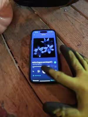 The image shows a phone on a wooden surface. The phone screen displays a music app interface with a song title and flower image on it. A hand with black-gloved fingers is interacting with the phone, touching the play/pause button. The wooden surface appears to be a table.
