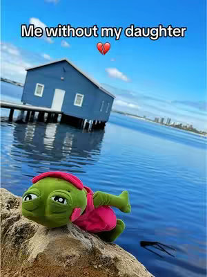 A plushie-like character in the foreground is placed on a rocky surface, positioned against a serene waterscape with a blue building on stilts and a clear sky. The plushie, featuring a green head, pink dress, and red headband, is the primary focal point. The water's surface reflects the sky, and the composition gives a sense of longing.