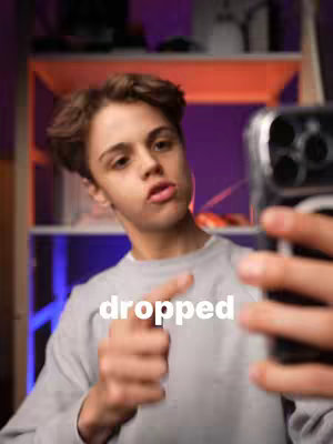 A young man in a gray sweater holds a phone up as if taking a selfie, pointing a finger in the foreground. White text reads "dropped".  The background has shelves with objects and purple/orange lighting.