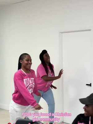A video cover featuring two young Black women dancing and smiling in a bright, clean room. They both wear pink tops and white or light blue pants, and are positioned to the left side of the frame, with one also wearing a headwrap. The focus is on their expressions and movement, suggesting happiness and energy. A person in a black cap can be partially seen in the bottom right corner of the frame. White walls and a partially visible door form the neutral background, placing the women in the foreground. Text is overlaid at the bottom, in the same color as the pink tops.