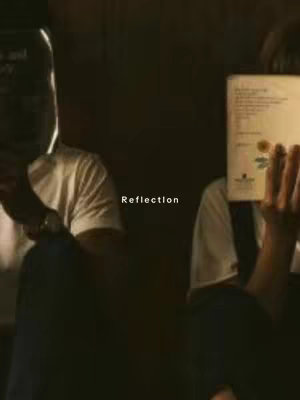 The image shows two people, partially visible, each holding a book. One person on the left is wearing a white shirt and holding a dark book obscuring their face, showing a watch. The second person on the right side is also wearing a white shirt and has a book in front of their face. The background is dark, and the overall lighting suggests a dim setting. In the center, the word 'Reflection' is written.