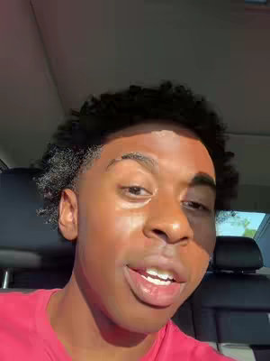 A young man with dark, textured hair and a slight smile is captured in a close-up shot. He's wearing a red shirt, and sunlight warmly illuminates his face, highlighting his features and casting shadows. The background suggests he might be in a car, with a car seat visible behind him, adding context to the selfie-style shot.
