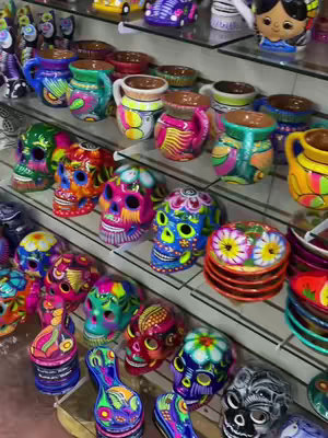The image is a vertical shot showcasing a display of vividly painted ceramic art. On glass shelves, a variety of items are presented: colorful mugs with handles, small bowls, and numerous decorative skulls. The mugs display a mix of bright colors and patterns, and the skulls are painted with intricate designs and bright colors. The overall composition is well-lit, showing the shiny glaze and detailed artwork.