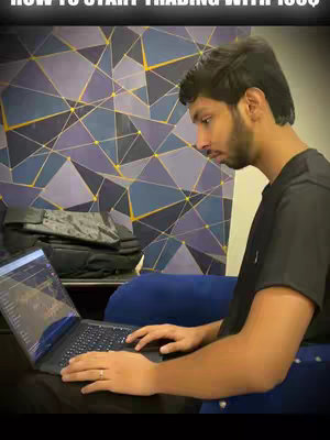 A man sits in a chair, using a laptop with a trading interface visible on the screen. The background features a patterned wall. The image has a warm, ambient light.