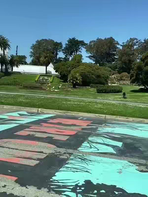 A view of a park with blue sky. In the foreground there is a road surface that has been painted with blue and red, in somewhat abstract shapes. Behind the road is a green area with grass and trees. In the distance there is more green space and a white building behind that.