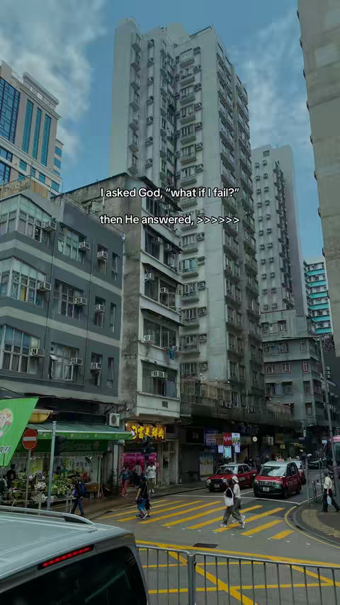 An urban street scene in Hong Kong, featuring tall apartment buildings, shops, pedestrians, and vehicles.