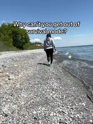 A woman walks away from the camera on a rocky beach toward the ocean. The sky is blue with some clouds. The text overlay says, 'Why can't you get out of survival mode?'