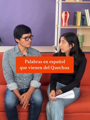 Two individuals sitting comfortably on an orange couch. In the center, a large, orange rectangular text overlay reads: 'Palabras en español, que vienen del Quechua' (Words in Spanish that come from Quechua). The background shows a white bookshelf with books and decorative objects, and a purple wall. The person on the left is wearing glasses and a patterned shirt, while the right person is wearing a black top.