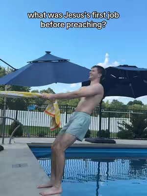 A man in swim trunks is seen leaning back, pulling on a yellow towel, near a pool. The background includes blue umbrellas, a fence, and a clear sky.