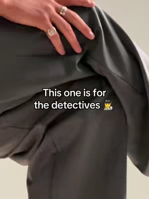 A close-up shot shows a person's arm and legs, clad in neutral-toned clothing. The focus is on the left arm, with several rings visible on the fingers. The hand rests on the upper thigh of the person's leg. Text is overlaid at the bottom: 'This one is for the detectives'.