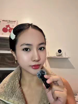 A young woman with dark hair styled with white clips is holding a small microphone. She has fair skin, red lipstick, and is wearing a brown fuzzy sweater. The background includes shelves and a sign featuring a cherry design.