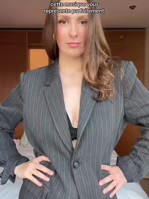 A woman in a gray pinstripe blazer stands in front of a blurred-out background. Her arms are at her sides, hands on her hips. Text overlays the top.