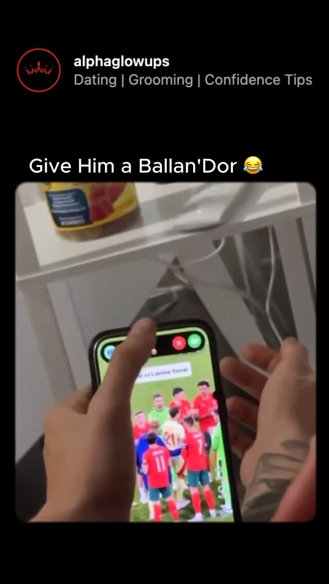 The image shows a person holding a phone, displaying a soccer match on its screen. The person's hands and a part of their arm are visible. Above, the text overlay reads, 'Give Him a Ballan'Dor 😂'. The phone screen shows a soccer field with players in red and green uniforms.