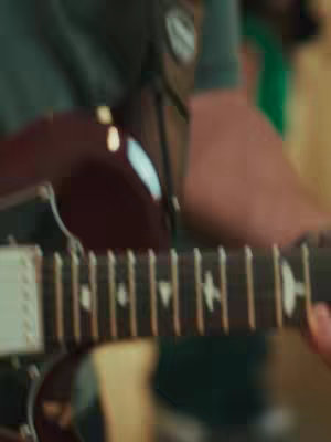 The image focuses on a guitar, showcasing a portion of the fretboard and strings. The composition is close-up, emphasizing the instrument's details. The color palette includes warm tones, with the wooden body of the guitar being the primary visual. The background slightly blurred to place more focus on the guitar itself.