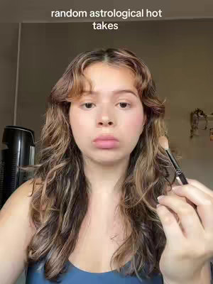 A medium shot shows a fair-skinned woman centered in the frame, looking directly at the camera with a neutral expression. She has wavy, light brown hair with darker highlights, styled with a side part and bangs. Her skin has a light makeup application with blush on her cheeks. She's holding a makeup brush near her face. The background is a plain, blurred shade of brown.