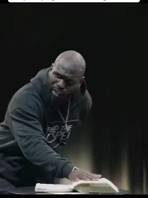 A man in a black hoodie stands behind an open book, possibly a bible. He is looking to the side, hand on the pages as if reading. The background is dark.