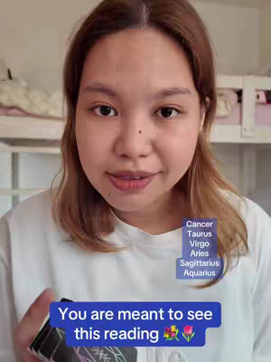 A woman is holding a deck of cards, looking at the viewer with a light smile. A blue banner at the bottom holds the message 'You are meant to see this reading' with a pair of flower and a tulip emojis. On the right side is a column of zodiac signs which are written in white. Behind her is a bed and a white wardrobe.