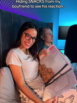 A woman with glasses and a man are seated on what appears to be a bed, a cookie-filled container held between them. The woman smiles at the camera, suggesting a positive or playful mood. The background suggests a domestic setting with minimal, modern decor. The woman wears a t-shirt and the man is behind her, implying an intimate setting.