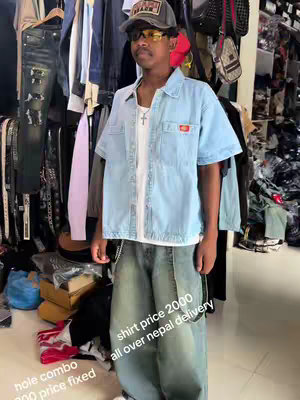 A young man stands centered, posing in a shop or closet filled with clothing racks and items. He wears a casual outfit: a light blue denim short-sleeved shirt over a white t-shirt and loose-fitting, acid-wash denim jeans. He's also wearing a grey baseball cap, sunglasses, and a silver chain. Text overlays the image with pricing and delivery information for the clothing.