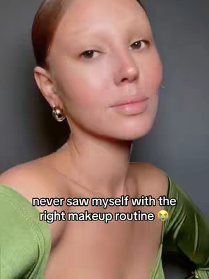 A close-up portrait of a person with a light complexion, fair eyebrows, and light eyes against a dark gray background. They wear a green, off-the-shoulder top and large gold hoop earrings. The face has a touch of makeup and a neutral expression. Text overlay is present at the bottom with a slight blush on the cheeks, the upper face is bare, and the individual appears to be looking slightly upwards and to their right. The lighting seems natural, and the overall composition is simple, focusing on the person's face.