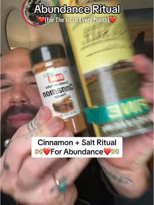 A man holds up a partially visible bottle of cinnamon and a container, the top of which says 'Abundance Ritual', written on the image. Text overlays read 'Cinnamon + Salt Ritual' and 'For Abundance'.  The background shows a partially visible ceiling with various colors