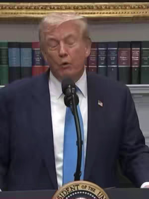 Image of Donald Trump speaking into a microphone behind a podium. He's wearing a dark suit, blue tie and a microphone in front of him. The background includes bookshelves.
