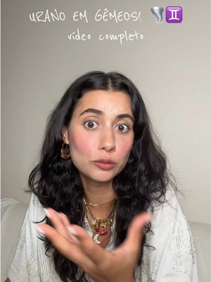 A woman with dark hair and wide eyes, wearing a white top and gold jewelry, extends her arm with an open palm as if presenting or explaining something. Text overlays the image indicating the video is about astrology relating to Uranus in Gemini.