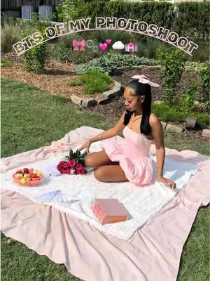 A young woman in a pink dress is sitting in a garden. She is on a picnic blanket with food and flowers. There is a text overlay that reads 'BTS OF MY PHOTOSHOOT'. The overall composition is well-lit and captures a moment of relaxation and celebration.