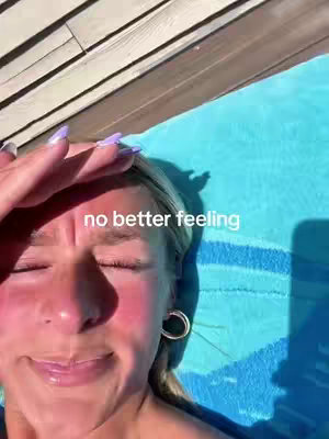 A close-up shot features a woman with fair skin, blonde hair, and a pair of hoop earrings. She is outside, as evidenced by the sunlight, with her eyes closed, and her hand is up towards her forehead as to block the sun. In the background are wooden boards and a blue towel. The text overlay reads, 'no better feeling'.