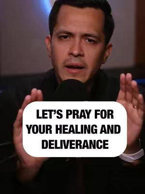 The image features a man in a close-up shot against a blurred background. The man is of mixed ethnicity, with short dark hair, wearing a dark-colored outfit. He is positioned centrally in the frame, with both hands raised, palms facing forward, as if in a gesture of prayer or supplication. A white-bordered text overlay is in the center of the frame with the message, 'LET'S PRAY FOR YOUR HEALING AND DELIVERANCE'. The lighting appears to be soft, highlighting his face. A microphone is placed in front of him.