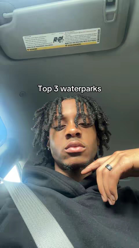 A selfie-style shot taken from inside a car. The main subject is a Black man with short dreadlocks, looking directly at the camera. The background features the car's interior, including the ceiling and sun visor. Text overlayed on the image reads 'Top 3 waterparks'. The man is wearing a black hoodie and a ring on his finger.