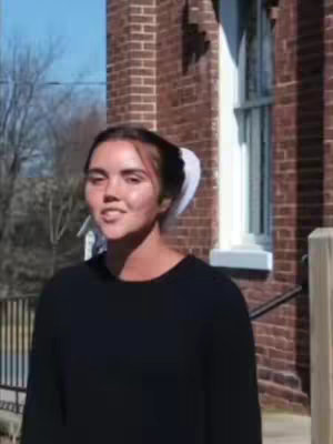 The cover image features a young woman positioned in front of a brick building with a large window. She is looking towards the camera, seemingly speaking. The lighting appears to be natural, as if taken outside on a sunny day.