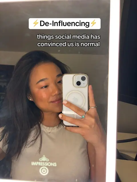A young woman is featured in a mirror selfie. She holds a phone up, slightly angled towards the mirror, which shows her face and upper body. The background shows a well-lit room and some clothes, reflecting off the mirror. Text overlays are added, along with a filter emphasizing the subject's skin tone.