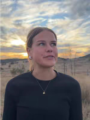 A woman with blonde hair and a black shirt stands outdoors, facing slightly upwards with a soft expression. The background shows a field with trees and a partly cloudy sky with a gold-toned sunset, indicating a time of day late afternoon or early evening.