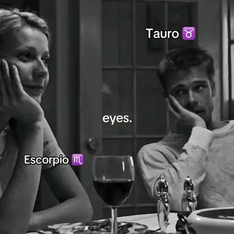 A black and white film scene featuring two people at a table, a woman, and a man. Text overlays identify them as zodiac signs. A glass of red wine is visible in the foreground.