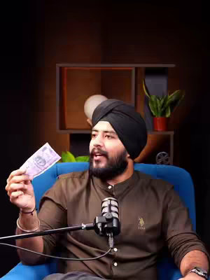 A man wearing a black turban, a brown shirt, and a trimmed beard is seated on a blue armchair. He's holding a purple Indian banknote in his left hand and a microphone in front of him. A background with some plant, a white sphere, and a dark wooden wall. The lighting is focused on the man and the microphone.