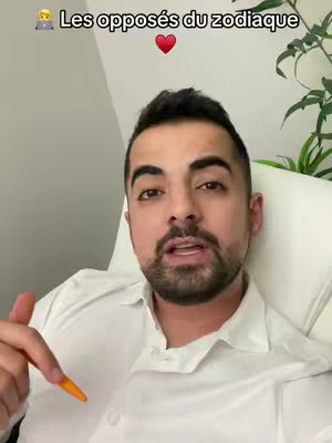 A man with dark hair and a beard is centered in the frame, speaking to the camera. He is wearing a white button-down shirt. Above the man's head, in the top center of the image, is the text "Les opposés du zodiaque" in a black, bold font. Below the text sits a red heart emoji. To the right of the man's head, a green plant is visible. The background appears to be a light gray wall. The image has a well-lit, studio-like quality.