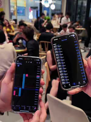 Two smartphones are held up, each displaying a trading chart, in front of a blurry background of people in a cafe setting.