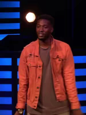 A man in an orange jacket stands in front of a blue panel backdrop with a single circular light above him.