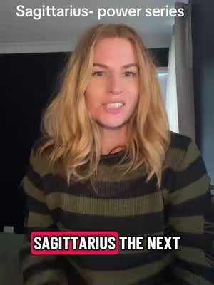 A woman with light-colored hair is in the foreground, speaking to the camera. Behind her is a neutral setting with a dark background and a light-colored curtain. The woman is wearing a green and black striped sweater, and the text overlay at the bottom of the video reads "SAGITTARIUS THE NEXT".